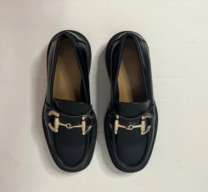 Svarta loafers med metallspänne H&M strl 35 - Snygga svarta loafers från H&M med chunky platt sula och dekorativt metallspänne framtill. Skorna har en rund tå och är tillverkade i syntetmaterial med en klassisk design som passar till många olika stilar. Aldrig använda då jag har oturen att ha små och för breda fötter..