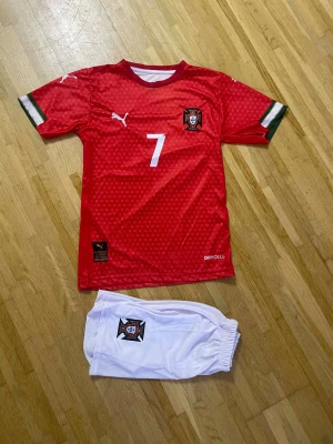 Portugal fotbollströja Ronaldo #7 - Säljer en röd Portugal fotbollströja med nummer 7 och Ronaldo på ryggen, samt vita shorts med Portugal-emblem. Tröjan har små mönster, gröna och vita detaljer på ärmarna och är tillverkad i polyester med DryCell-teknologi. Perfekt för dig som älskar fotboll och Ronaldo.