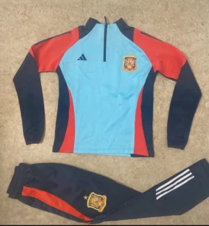 Spaniens Adidas fotbollsträningsoverall - Säljer en officiell träningsoverall från Spaniens landslag med Adidas-logga. Tröjan är ljusblå med röda och mörkblå detaljer, half zip och landslagsemblem på bröstet. Byxorna är mörkblå med röda inslag, vita ränder och landslagsemblem på benet. Perfekt för fotbollsträning.