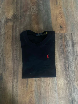 Svart t-shirt från Polo Ralph Lauren - Klassisk svart t-shirt från Polo Ralph Lauren med röd broderad logga på bröstet. T-shirten har rund halsringning och korta ärmar. Tillverkad i mjuk bomull för skön känsla och enkel stil.