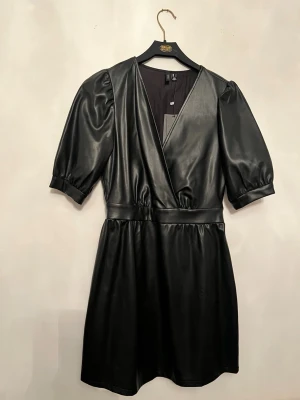 Svart klänning i skinnimitation - Snygg svart klänning från VERO MODA  i skinnimitation med puffiga korta ärmar och markerad midja. Klänningen har en v-ringad front och är tillverkad i syntetmaterial som ger en cool läderlook. Perfekt för dig som gillar edgy stil och vill sticka ut. Det finns lite defekt på klänningen som ni kan se på sista bilden men inget som man märker av. 