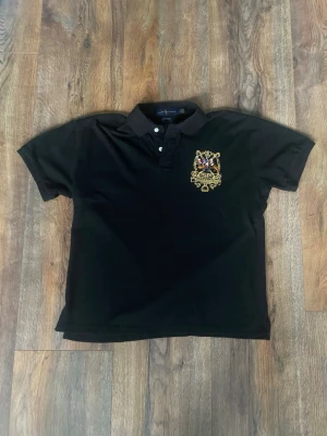 Svart piké från Ralph Lauren - Säljer en svart pikétröja från Ralph Lauren med broderat polomotiv och guldfärgade detaljer på bröstet. Tröjan har korta ärmar, krage med knappar och en röd siffra 3 på ena ärmen. Materialet är mjuk bomull och passformen är normal. Storlek är INTE L som det står på den. Den är liten i storlek och passar S.