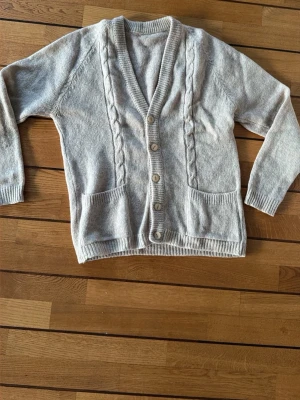 Grå stickad kofta med knappar - Mysig grå stickad kofta med kabelstickade detaljer längs fram och fem träknappar. Koftan har två praktiska fickor och ribbade muddar vid ärmslut och nederkant. Perfekt för lager-på-lager under kyliga dagar.