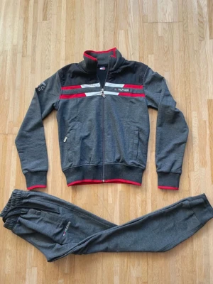 Grå mjukisbyxor från Tommy Hilfiger - Snygga grå mjukisbyxor från Tommy Hilfiger med broderad logga på benet. Byxorna har elastisk midja med snörning och dragkedjeficka på sidan. Materialet är mjukt och bekvämt, perfekt för chill eller träning. Diskreta röda detaljer och slim passform.