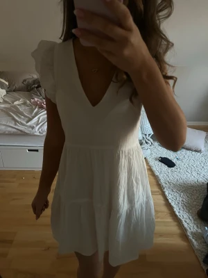 Vit kortklänning med volangärmar - Supersöt vit klänning med v-ringning och volangdetaljer på axlarna. Klänningen är kort och har en luftig, lätt struktur i bomull. Perfekt för varma dagar och har en feminin siluett. Klänningen har dubbelt tyg vid kjolen. Materialet är mjukt och känns skönt mot huden. Klänningen är endast använd en gång, till min konfirmation. Den har inga defekter.