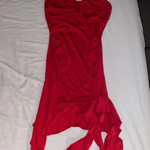 Röd axelbandslös klänning - Säljer en röd axelbandslös klänning med asymmetrisk nederkant och draperade detaljer. Klänningen har en snygg, figurnära passform och är perfekt för dig som vill sticka ut. Materialet känns mjukt och följsamt mot kroppen.