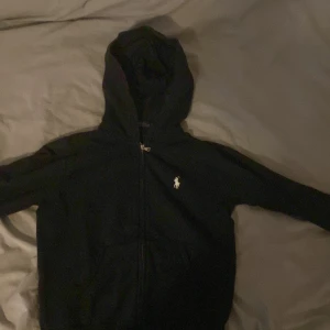 Svart hoodie från Polo Ralph Lauren - Svart hoodie från Polo Ralph Lauren med dragkedja och klassisk vit logga på bröstet. Säljer denna för att den är alldeles för liten för mig, bra skick men dragkedjan är lite sliten