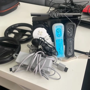 Nintendo Wii Konsol med Tillbehör - Nintendo Wii i svart med tre Wii Remote-kontroller (vit, blå, svart), två rattar, nunchuk, strömadapter och sensorbar. Allt ser ut att vara i gott skick med mindre ytslitage. Perfekt för familjespel och partykvällar!