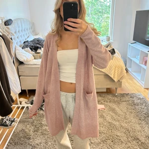 Ljusrosa lång kofta med fickor - Supermysig ljusrosa lång kofta med två stora fickor framtill och ribbade muddar. Koftan har en relaxed passform och är perfekt att slänga över en topp för en chill look. Materialet känns mjukt och skönt mot huden.