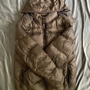 Brun pufferjacka från Calvin Klein - Säljer en brun pufferjacka från Calvin Klein Jeans med avtagbar huva och dragkedja framtill. Jackan har två fickor med dragkedja och ett stort CK-logomärke på ärmen. Fodret är mönstrat med CK-loggor för en extra snygg touch.