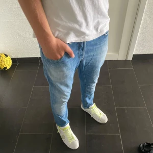 Dondup George - Säljer ett par ljusblå jeans från Diesel med Dondup-detalj på bakfickan och klassisk orange söm. Jeansen har normal passform och raka ben, tillverkade i mjukt denimtyg. Snygga detaljer som metall-D på bakfickan och röd kontrastsöm vid loggan.