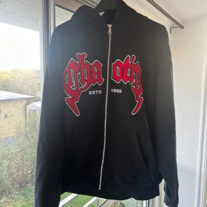 Svart hoodie med röd tryck från FSBN - Svart hoodie från FSBN i relaxed fit med dragkedja och huva. Framsidan har ett stort tryck i rött och vitt med texten 'ESTD. 1992'. Mjuk insida och två fickor framtill. Perfekt för dig som gillar streetwear vibes.