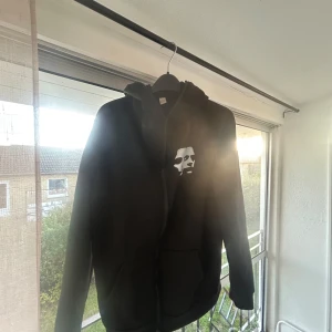 Svart hoodie med vit ansiktsgrafik - Svart hoodie med stor vit ansiktsgrafik tryckt på ryggen och mindre tryck på bröstet. Tröjan har huva, långa ärmar och känguruficka framtill. Perfekt för dig som gillar streetstyle och vill sticka ut.