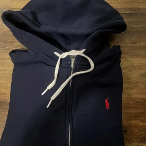 Marinblå hoodie från Polo Ralph Lauren - Marinblå hoodie från Polo Ralph Lauren med vit snörning och dragkedja framtill. Klassisk röd logga broderad på bröstet och stora fickor. 