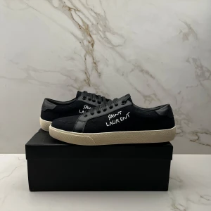 Svarta Saint Laurent Skor - Svarta skor från Saint Laurent med vit handskriven logga på sidan. Skorna har svart ovandel i canvas och skinn, svarta snören och en kontrasterande vit sula. Klassisk låg modell med rund tå och diskreta detaljer. Perfekta för en stilren och trendig look.