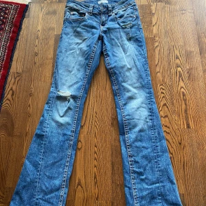 Blå bootcut jeans med slitningar - Snygga blå jeans med bootcut passform och låg midja. Jeansen har slitningar på låret och detaljerade bakfickor med knappar. Klassisk femficksmodell i jeansmaterial, perfekt för en avslappnad och trendig look.