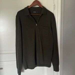 Mörkgrön stickad tröja J.Lindeberg - Snygg mörkgrön stickad tröja från J.Lindeberg med krage, bröstficka och half zip-dragkedja. Perfekt för dig som gillar stilrena plagg med en twist. Långärmad och mjuk, passar till många olika outfits.