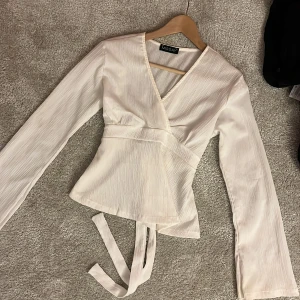 Vit omlottblus från Missay - Säljer en stilren vit omlottblus från Missay med långa ärmar och knytband i midjan. Blusen har v-ringning och är tillverkad i ett lätt, strukturerat tyg som ger en snygg och modern look. Perfekt att styla med jeans eller kjol för en trendig outfit.