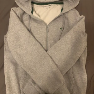 Grå hoodie från Lacoste med dragkedja - Snygg grå zip hoodie från Lacoste i topp skick och utan några defekter.