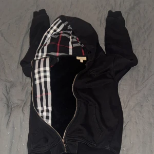 Svart Burberry hoodie med rutig huva - Svart hoodie från Burberry med dragkedja och klassiskt rutig foder i huvan. Broderad Burberry-logga på bröstet. Mjuk insida och stora fickor framtill. Perfekt för dig som vill ha en stilren och lyxig streetwear-look.