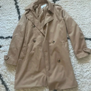Beige trenchcoat med bälte vero moda brun  - Snygg beige trenchcoat med klassisk krage, dubbelknäppning och bälte i midjan. Jackan har långa ärmar med knäppning vid ärmslut och två sidfickor. Perfekt för dig som vill ha en stilren och tidlös look. s passar m 