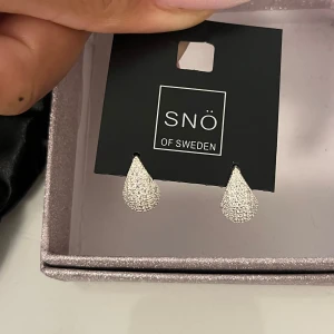 Glittriga droppformade örhängen SNÖ - Snygga droppformade örhängen från SNÖ of Sweden i silverfärg med glittrig yta. Perfekta för att ge din look extra bling! Modellen heter Naomi drop ear s/clear och de kommer i original förpackning, aldrig andvända❤️original pris 349kr