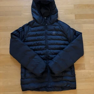 Svart pufferjacka från Adidas XS - Svart pufferjacka från Adidas med huva och klassiska tre ränder på ärmarna. Jackan har dragkedja framtill och är quiltad med horisontella sömmar. Perfekt för kyliga dagar och har en sportig vibe.