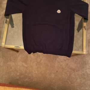 Mörkblå Moncler stickad tröja - Snygg mörkblå stickad tröja från Moncler med klassisk rund hals och diskret logga på bröstet. Tröjan har ribbade muddar och en stilren look med röd och vit rand på insidan av kragen. Perfekt för dig som gillar exklusiva och tidlösa plagg.