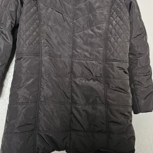 Svart quiltad pufferjacka Janet.Y 2XL - Svart pufferjacka från Janet.Y i storlek 2XL med quiltade detaljer på axlar och sidor. Jackan har långa ärmar, knappar framtill och två fickor med dragkedja och dekorativa knappar. Perfekt för kalla dagar med sin snygga, lite oversized look.