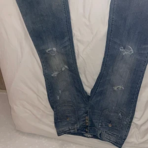 Blå jeans med slitningar och knappar - Säljer ett par blå jeans med slitna detaljer och snygga knappar framtill. Jeansen har stora fickor med lock och knapp både fram och bak, samt dekorativa sömmar. Perfekt för dig som gillar en avslappnad och cool stil. Det står ingen exakt storlek men skulle gissa på de passer en 42-44