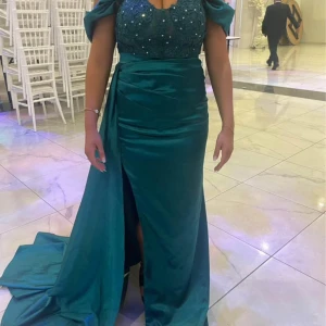 Mörkgrön satin klänning  - Säljer en mörkgrön långklänning i glansigt satinliknande material. Klänningen har offshoulder-ärmar, pärldetaljer på överdelen och en snygg slits framtill. Kjolen har ett elegant släp och överdelen är dekorerad med små pärlor för extra glam.