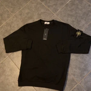 Svart stone island sweatshirt - Svart långärmad tröja från Stone Island med klassisk rund hals och ikonisk logotyp-patch på vänster ärm. Tröjan är i mjuk bomull och har en clean, stilren look som passar perfekt till streetwear. Ribbstickade muddar vid ärmslut och nederkant.