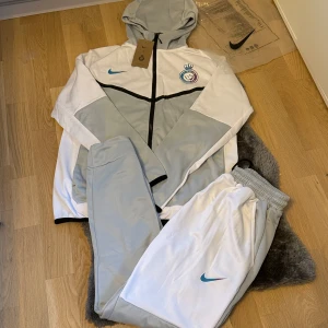 Nike tech set/tracksuit al nassar - Nike hoodie och byxor i vitt och ljusgrått med svart dragkedja och huva. Tröjan har klubbmärke på bröstet och blå Nike-logga. Perfekt för träning eller chill, med sportig look och bekväm passform.