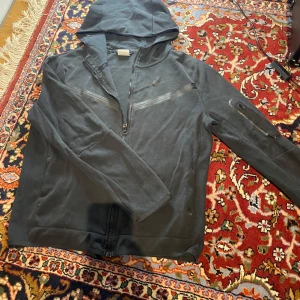 Svart Nike Tech  hoodie med dragkedja (äkta)(Nike tech passar både män och kvinnor) - Svart hoodie från Nike (äkta) med hel dragkedja och huva. Snygga svarta detaljer i form av ett diskret Nike-logga och ett band över bröstet. Långärmad med ficka på ärmen och mjukt bomullsmaterial. Perfekt för chill eller träning. Diskutera gärna pris! 😊😎Nypris: 950 kr!😊 Nike tech passar både män och kvinnor