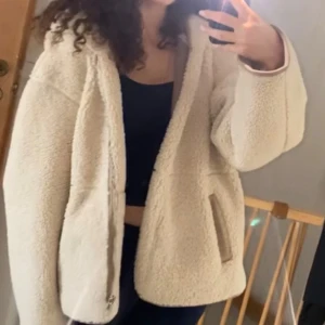 Beige teddyjacka - Mysig beige teddyjacka med bruna detaljer, huva och dragkedja framtill. Jackan har en loose passform, två fickor och är tillverkad i fluffigt fleece-material. Perfekt för kalla dagar när du vill vara både varm och stylish. 