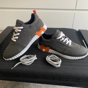 Skor - Snygga grå sneakers med vit räfflad sula och coola orange detaljer på sidan och hälen. Skorna har vit snörning och är tillverkade i skinn med en modern, sportig look. Perfekt för dig som gillar exklusiva märken och unik design. Låda medföljer tyvärr inte!
