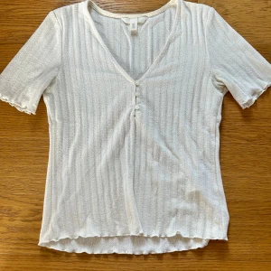 Vit ribbad topp med knappar från H&M - Säljer en vit ribbad topp från H&M i storlek S. Toppen har v-ringning med tre små knappar framtill, korta ärmar och vågiga kanter vid ärmslut och nederkant. Pris går att diskuteras!