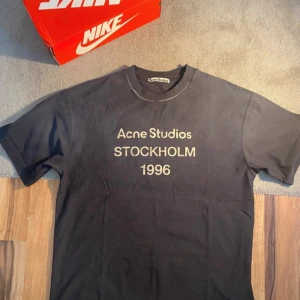 Svart Acne Studios t-shirt Stockholm - Svart t-shirt från Acne Studios med tryck 'STOCKHOLM 1996' i vitt på bröstet. Klassisk rund hals och korta ärmar. Materialet är mjuk bomull och passformen är normal. Perfekt för dig som gillar stilrena och ikoniska plagg.