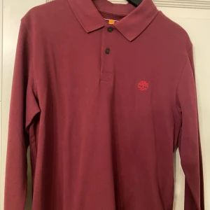 Vinröd långärmad pikétröja Timberland - Säljer en vinröd långärmad pikétröja från Timberland i slim fit. Tröjan har klassisk krage, två knappar och broderad logga på bröstet. Tillverkad i 100% ekologisk bomull för en skön känsla och snygg look.