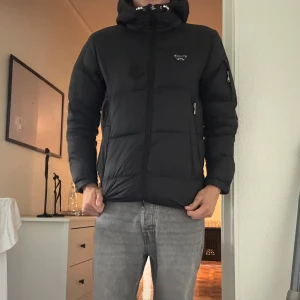 Svart alessandro zavetti pufferjacka med huva - Svart pufferjacka med huva och dragkedja framtill. Jackan har quiltad design, flera fickor med dragkedja och en liten logga på bröstet. Perfekt för kalla dagar och har en klassisk, stilren look. Sen är sett ett hål i jackan som är lagat annars är det skön varm och snygg storlek m passar även L ställ frågor om ni har några