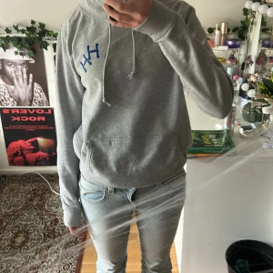 Grå hoodie Just Hoods Känn Ingen Sorg. (Håkan Hellström) - Grå hoodie från Just Hoods med blått tryck 'KÄNN INGEN SORG' på ryggen och blå HH-logga på bröstet. Klassisk känguruficka, snörning i huvan och mjukt material i bomullsblandning. Perfekt för dig som gillar statement-plagg och chill stil.
