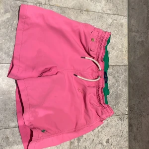Rosa badshorts från Polo Ralph Lauren - Snygga rosa badshorts från Polo Ralph Lauren med grön midja och klassisk broderad logga i grönt. Shortsen har snörning i midjan och en liten ficka med knapp. Perfekta för stranden eller poolen.