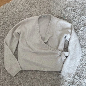 Beige omlott kofta från Kappahl - Säljer en mjuk och mysig beige omlott kofta från Kappahl med lång ärm och ribbade muddar. Koftan har v-ringad hals och knyt i sidan för en snygg passform. Perfekt att slänga på sig när det är lite kyligt ute.💕