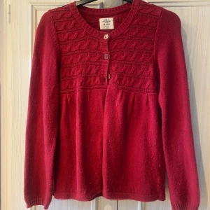 Röd kofta med kabelstickat mönster - Mysig röd kofta från H&M med kabelstickat mönster upptill och rund halsringning. Koftan har tre knappar framtill och långa ärmar. Perfekt för lager-på-lager och ger en klassisk look till din outfit. På lappen står det att den är storlek 146-152 men jag som har S kan ha den 