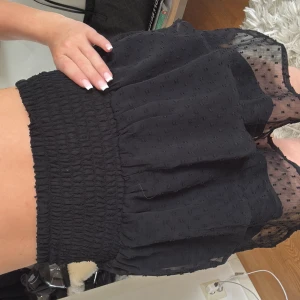 Svart volangkjol med transparenta detaljer - Supersöt svart kortkjol med volanger och transparenta partier i mesh med små prickar. Kjolen har bred smock i midjan för en bekväm passform och ett lekfullt lager-på-lager-utseende. Perfekt för dig som gillar trendiga och feminina plagg.