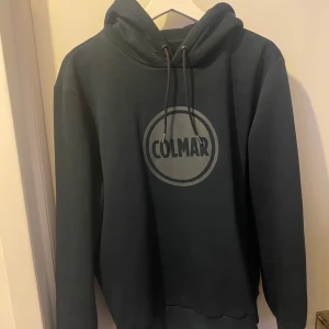 Blå hoodie från Colmar med logga - Säljer en snygg marinblå hoodie från Colmar med stort 3D-logotryck på bröstet. Tröjan har huva med snörning och ribbade muddar vid ärmslut och nederkant. Perfekt för dig som gillar streetwear och vill ha en stilren look.