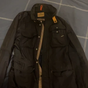 Svart vindjacka Parajumpers M - Svart vindjacka från Parajumpers med flera praktiska fickor, dragkedja och knappar framtill. Jackan har coola orange detaljer och meshfoder på insidan för extra ventilation. Perfekt för dig som vill ha en funktionell och snygg jacka med streetkänsla.