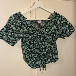 Blommig blus med snörning från Lindex - Supersöt blommig blus från Lindex i svart med grönt och vitt blommönster. Toppen har korta ärmar, v-ringning och snörning framtill som ger en snygg detalj. Perfekt för dig som gillar en romantisk och trendig stil. Passar perfekt till alla slags tillfällen. Från fest till vardag 