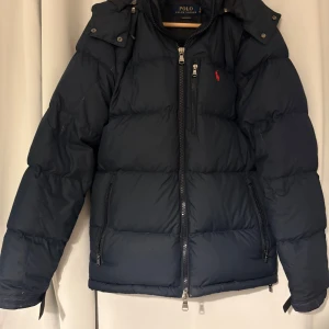 Mörkblå dunjacka Polo Ralph Lauren XS - Ord. Pris 6000kr!! Snygg mörkblå dunjacka från Polo Ralph Lauren med huva och flera dragkedjefickor. Jackan har en klassisk pufferstil, diskret logga på bröstet och är perfekt för kalla dagar. Passformen är normal och materialet är polyester.