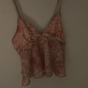 Blommig volang-linnetopp i pastell - Supersöt linnetopp med blommigt mönster i rosa, gul och röd pastell. Toppen har tunna axelband, volangdetaljer och är lätt transparent. Perfekt för varma dagar och ger en romantisk vibe med sitt luftiga tyg och v-ringning.